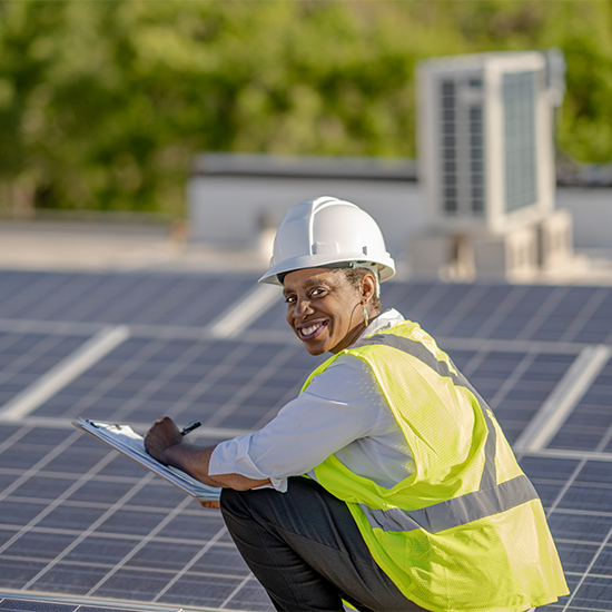 Smiling employee wearing a hard hat kneeling next to solar panels writing on a notepad